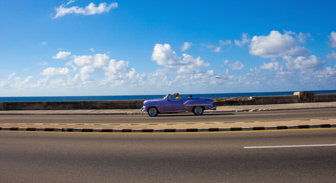 Havana, Cuba, a shiny purple convertible classic car on an empty road with water and clouds in the background