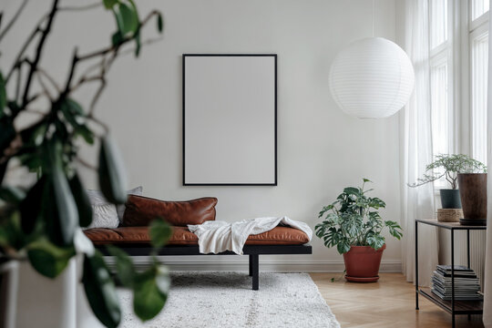 Frame mockup on a white wall in scandinavian natural style living room, brown leather ottoman, potted plants. Empty blank vertical din A poster with black frame in light room interior design