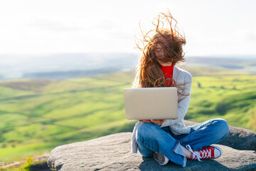 Woman Working on Laptop Outdoors on Rocky Outcrop Amid Rolling Green Hills in Spring