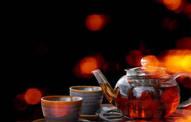 Glass teapot with tea, cups in close-up on a dark background with a bokeh effect.