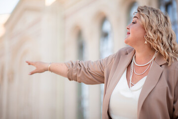 A beautiful fat middle aged woman walks around the city in a stylish image. The plus size model is dressed in a biege jacket and a white dress