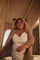 Fototapeta premium A beautiful fat middle aged woman walks around the city in a stylish image. The plus size model is dressed in a biege jacket and a white dress