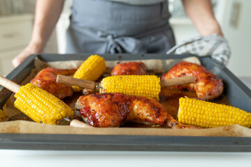 Woman serving delicious homemade food with oven baked and glazed chicken legs with corn on the cop on a baking tray