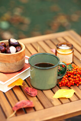 Cozy autumn scene with coffee, chestnuts, a book, and a candle on a wooden table, surrounded by fallen leaves and berries.