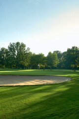 Tranquil Golf Course with Sand Bunker and Flag surrounded by lush trees under a clear blue sky. Perfect for concepts of relaxation, nature, and outdoor sports.