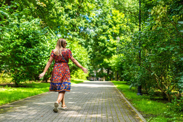 Beautiful blonde woman wearing colorful floral flowing dress walking against green background in city park on sunny day. Back view