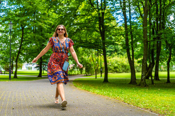 Beautiful blonde woman wearing colorful floral flowing dress walking against green background in city park on sunny day. Front view