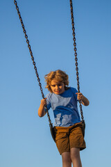 Kid swinging on swing in city park. Childhood, Freedom, Happy, Summer Outdoor. Cute child boy playing on swing, having fun. Blonde cute kid having fun on a swing in playground kids.