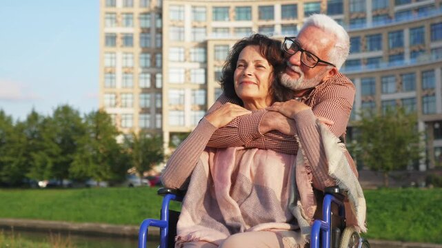 Senior woman in wheelchair walking with caregiver old man on road in park. Elderly family couple man supporting embracing paralyzed woman in chair for people with disability outdoor. Rehabilitation