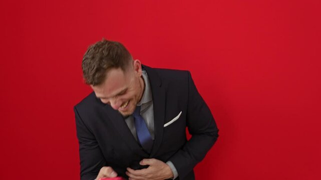 Young hispanic man in suit, laughing out loud while having fun with a funny joke on his smartphone, smiling against an isolated red background