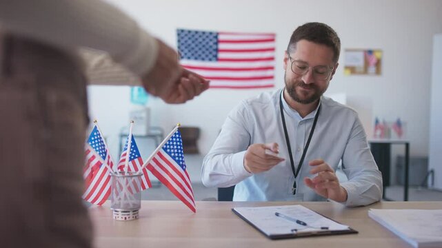 Smart Caucasian male with glasses sitting at desk. Accepting ID from someone behind camera and checking information on card. Miniature American flags located on table. People voting for president.