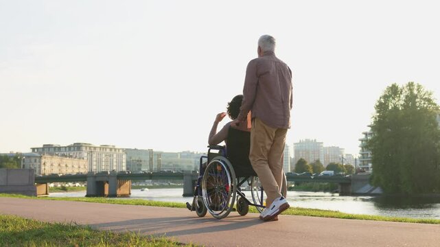 Senior woman in wheelchair walking with caregiver old man on road. Family couple man husband supporting taking care of paralyzed woman wife in chair for people with disability outdoor. Rehabilitation