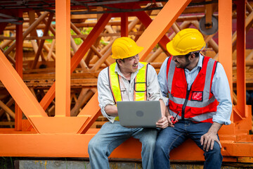 Two men in safety gear are looking at a piece of machinery. They are wearing reflective vests and...