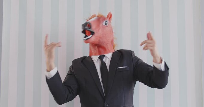 Cheerful eccentric businessman in black suit, wearing red horse rubber mask, snapping his fingers while dancing with his hands up. Dancer in bizarre carnival horse head on vertical striped background