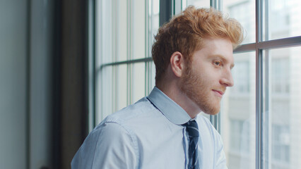 Fototapeta premium Head And Shoulders Portrait Of Smiling Young Businessman Looking Out Of Window In Office