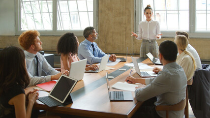 Young Businesswoman Giving Presentation To Multi-Cultural Team Sitting Around Table In Modern Office