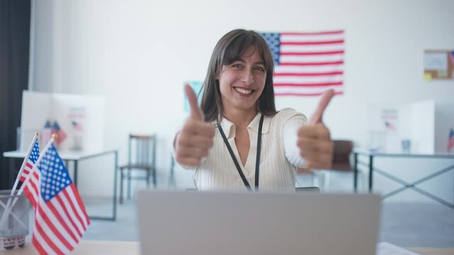 Happy girl sitting in office or polling station with American flags. Widely smiling and showing thumbs up to camera. Enjoying end of presidential elections. Motivating people to visit voting location.