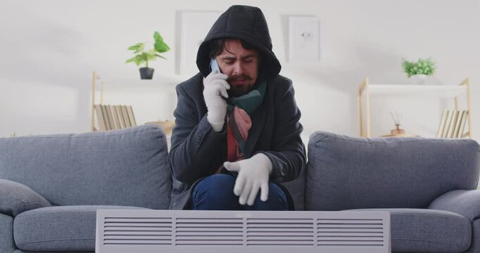 Young guy freezing at home with heating problem in winter. Man in hooded coat talking to heating service operator in living room in winter. Heating problems in cold season in winter.