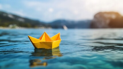 A yellow paper boat floats gently on a tranquil body of water. The water is clear and blue, reflecting the sunlight. In the background, a mountain range rises up against a hazy blue sky.