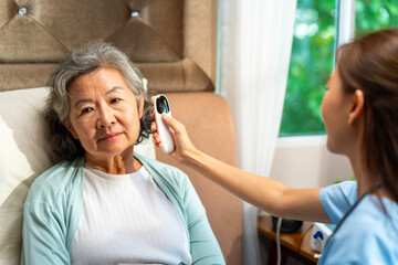 Asian woman doctor examine patient use infrared thermometer measured temperature in senior woman ear in bedroom. Nurse visit and checking elderly patient health at home. Home medical therapy concept.