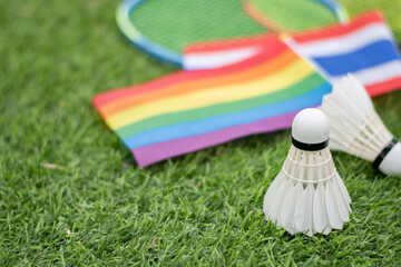 White cream shuttlecock, badminton sport equipment, places on green grass in front of Thailand national flag and rainbow flag, concept for badminton sport for all genders and lgbtq+ people.