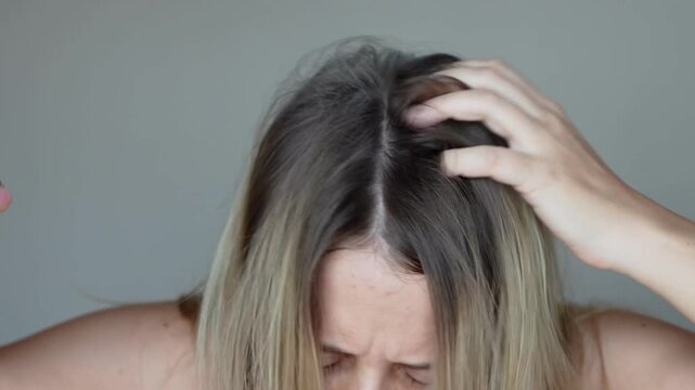 Young caucasian blonde woman with dirty greasy hair spraying dry shampoo on the roots of her hair rubbing it into the scalp with hands on a light grey background. A quick way to cleanse