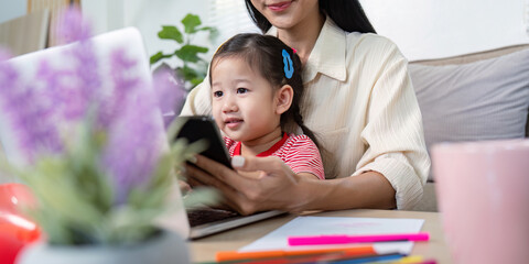Obraz premium Mother Working from Home with Toddler on Lap Using Laptop and Smartphone in Cozy Living Room