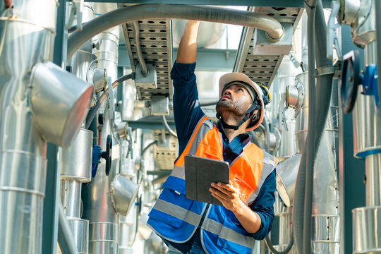 Professional Asian man engineer in safety uniform working on digital tablet at outdoor construction site rooftop. Industrial technician worker maintenance checking building exterior air HVAC systems.