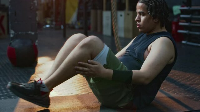 Tired young sportsman pumping abdominals flexing legs on training mat after gym workout