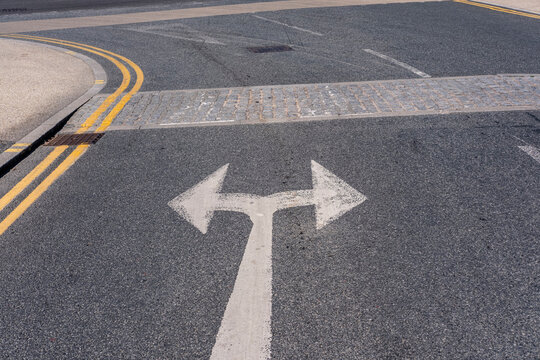 A close-up of a road surface with bold white arrows marking directions for left and right turns, accompanied by yellow lines and a paved crosswalk in a city setting.