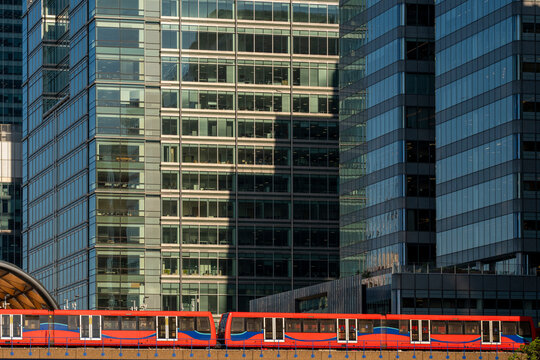A modern red train passing through a cityscape filled with tall glass buildings, showcasing urban architecture and public transportation during the daytime.