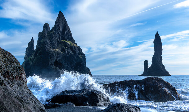Reynisdrangar spires at Reynisfjara Black Sand Beach, Iceland. A rocky shoreline with a large wave crashing into the rocks