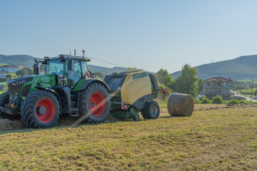 A green tractor picks up and shapes a bale of hay. The tractor is surrounded by a field of grass.
