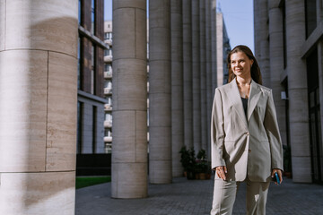 Confident businesswoman walking between modern office buildings, wearing a sleek suit. She exudes professionalism and success in an urban corporate environment