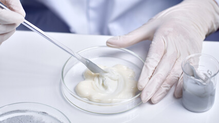 Cosmetic research in laboratory scene was shot from macro above view, the scientist holding a glass petri dish placed on white table and using glass rod to stir white cream above. Advertising space