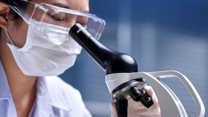 Female scientist in sterile clothing and safety goggles using a microscope to conduct research investigations in laboratory. Close-up from side view photograph for advertising pic designing