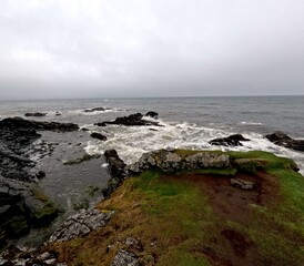 Misty Icelandic coastline with waves crashing on a rocky shore. The contrast of green grass and dark rocks highlights the dramatic beauty of the North Atlantic.