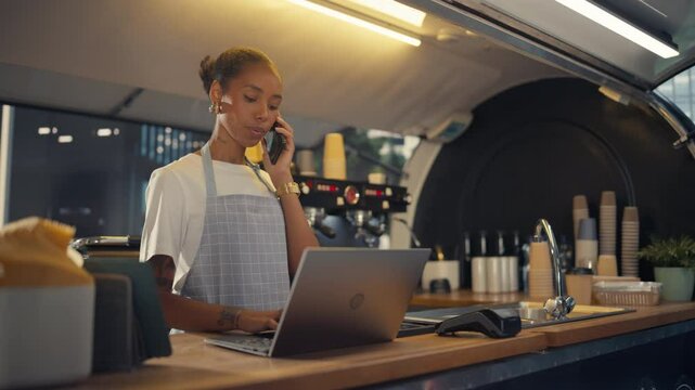Street Cafe Manager Having a Conversation on a Phone and Working on a Laptop Computer Behind a Counter. Black Female Entrepreneur Ordering Supplies Online, Managing Catering Menu