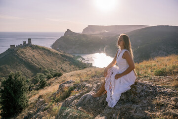 A woman in a white dress sits on a rock overlooking a body of water. The scene is serene and peaceful, with the woman enjoying the view and the calmness of the surroundings.