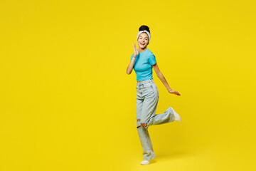 Full body side view young Latin woman she wearing blue top t-shirt headband casual clothes standing looking camera put hand on face raise up leg isolated on plain yellow background. Lifestyle concept.