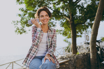 Young fun woman wear shirt casual clothes listen recorded voice message on mobile cell phone sits walk rest relax in spring sunshine green city park near sea outdoors on nature. Urban leisure concept.