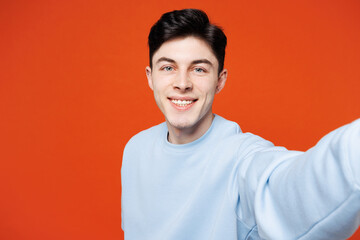 Close up young happy smiling man he wearing blue sweatshirt casual clothes doing selfie shot pov on mobile cell phone isolated on plain red orange color background studio portrait. Lifestyle concept.