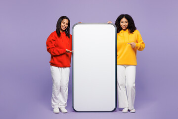 Full body two young little kid teen girl women they wear red yellow hoodie casual clothes point finger on big huge blank screen mobile cell phone isolated on plain purple background Lifestyle concept