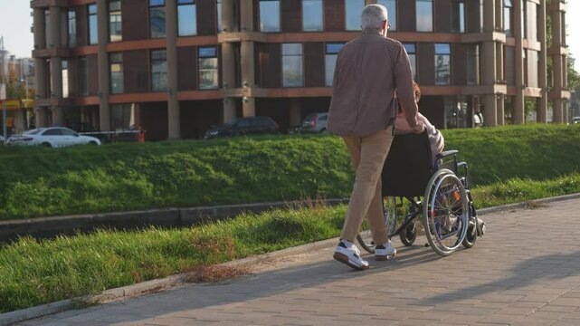Senior woman in wheelchair walking with caregiver old man on road. Family couple man husband supporting taking care of paralyzed woman wife in chair for people with disability outdoor. Rehabilitation