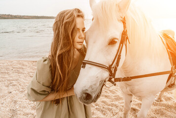 A woman in a dress stands next to a white horse on a beach, with the blue sky and sea in the...