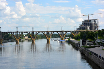 Obraz premium Beautiful view of the wide and long blue Dnipro River. Landscapes, a gray brick embankment with green trees, new construction sites and a beautiful bridge in the city of Dnipro.