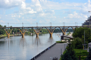 Beautiful view of the wide and long blue Dnipro River. Landscapes, a gray brick embankment with green trees, new construction sites and a beautiful bridge in the city of Dnipro.