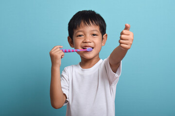 A happy Asian boy with clean teeth brushing his teeth while giving a thumbs up to the camera. Isolated on blue background. Health care, oral hygiene