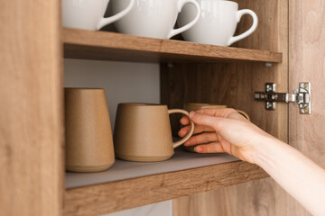 A person selecting a brown ceramic mug from a wooden cabinet filled with white coffee cups