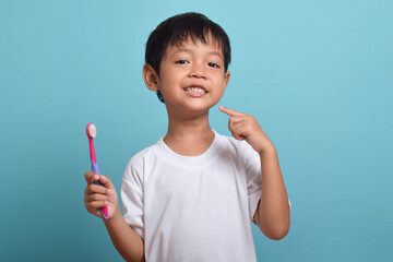A happy boy is holding a toothbrush while pointing at his teeth. Isolated on a blue background. The boy smiles showing his teeth. Children's dental care and hygiene